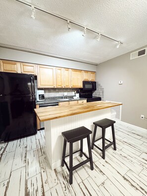 A modern kitchen with wooden cabinets, a black refrigerator, a microwave, and a wooden island with two stools.