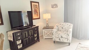 Una sala de estar con un televisor sobre un mueble negro, un sillón estampado, una mesita con una lámpara y una decoración en la pared.