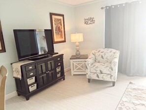 A living room with a TV on a black cabinet, a patterned armchair, a small table with a lamp, and a wall decoration.
