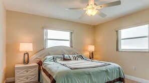 A bedroom with a bed, two bedside lamps, a ceiling fan, and two windows.