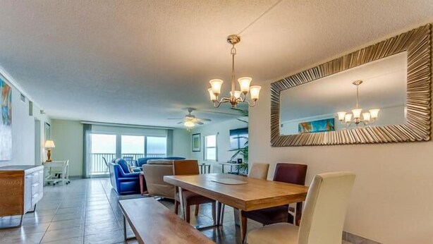 A spacious dining area with a large wooden table, multiple chairs, and a chandelier. There is a balcony visible in the background.