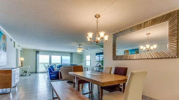 A spacious dining area with a large wooden table, multiple chairs, and a chandelier. There is a balcony visible in the background.