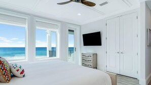 Une chambre dotée d’un grand lit, d’un ventilateur de plafond, d’un téléviseur fixé au mur et offrant une vue sur l’océan grâce à de grandes fenêtres.