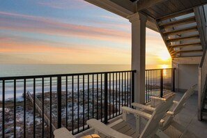 En balkon med udsigt over havet i solnedgang, med træterrasse og to Adirondack-stole.