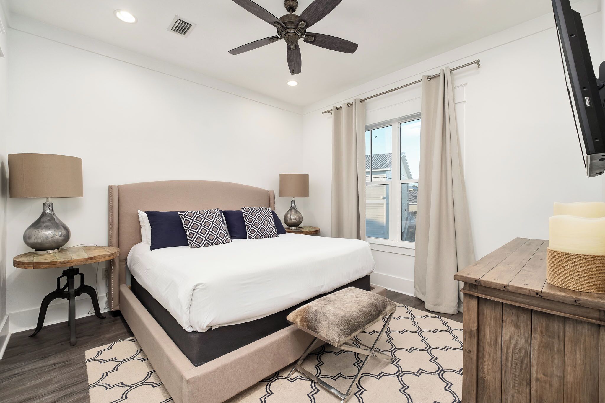 A bedroom with a bed, two bedside tables, a ceiling fan, a window with curtains, and a television.