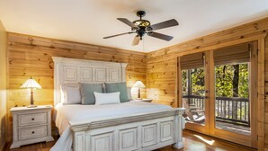A bedroom with a wooden ceiling, a bed with white bedding, two bedside tables with lamps, and a view of the outdoors through a glass door. - Log Cabin, Lake, Hot Tub, Near Wineries & Hiking (Dahlonega)