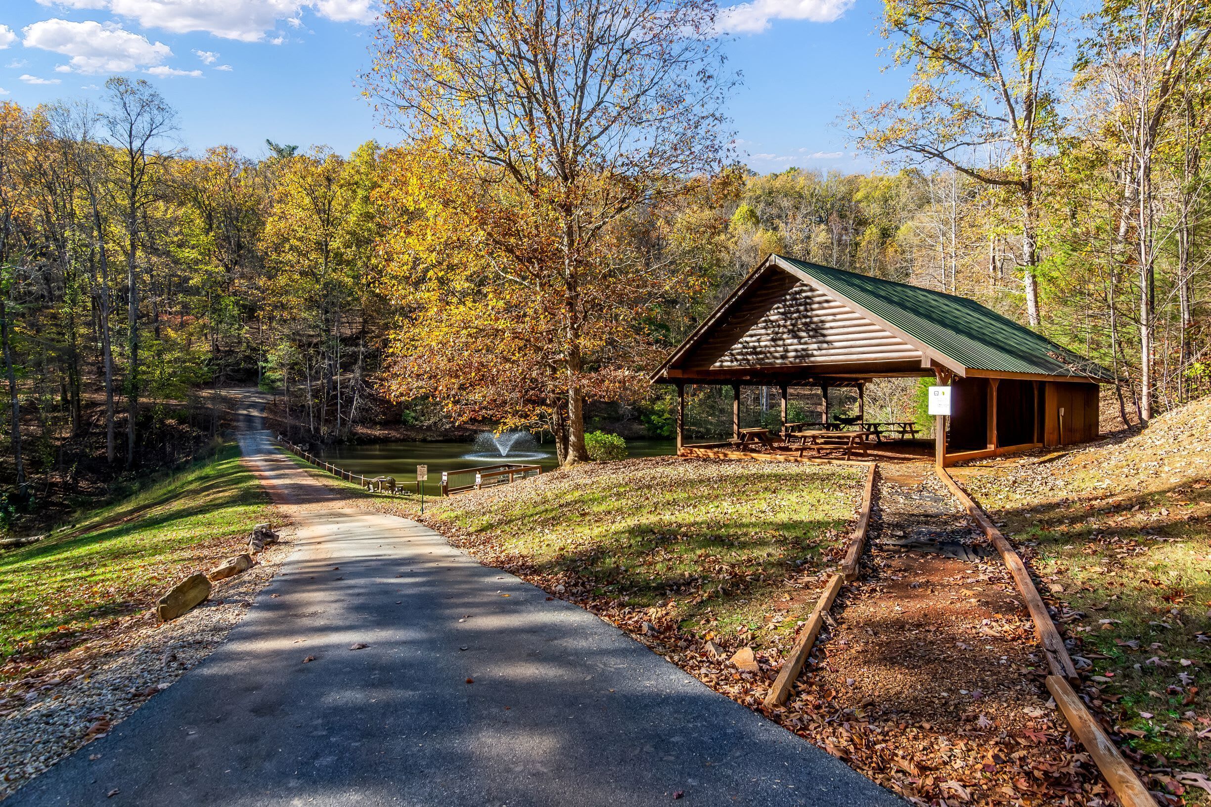A covered picnic area with a picnic table and benches, surrounded by a forested area with a paved path and a small fountain.