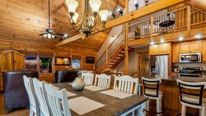 A spacious wooden cabin interior with a dining area, kitchen, and a staircase leading to a loft. - Log Cabin, Lake, Hot Tub, Near Wineries & Hiking (Dahlonega)