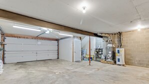 Un garage avec un sol en béton, une porte de garage blanche, un éclairage fluorescent et un chauffe-eau adossé à un mur en briques.