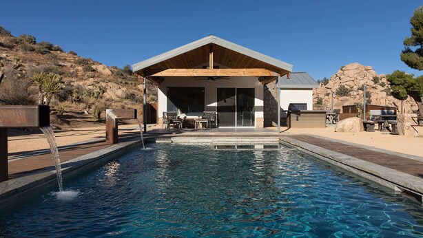 Un área de piscina moderna al aire libre con una zona de estar techada, una fuente de agua y montañas al fondo.