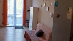 Ett rum med en säng, ett kylskåp, ett fönster med gardiner, en dörr och en väggmonterad ljusströmbrytare.