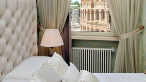 Ein Schlafzimmer mit einer gepolsterten Kopfteil, einer Nachttischlampe und Blick auf das Kolosseum durch Vorhänge.