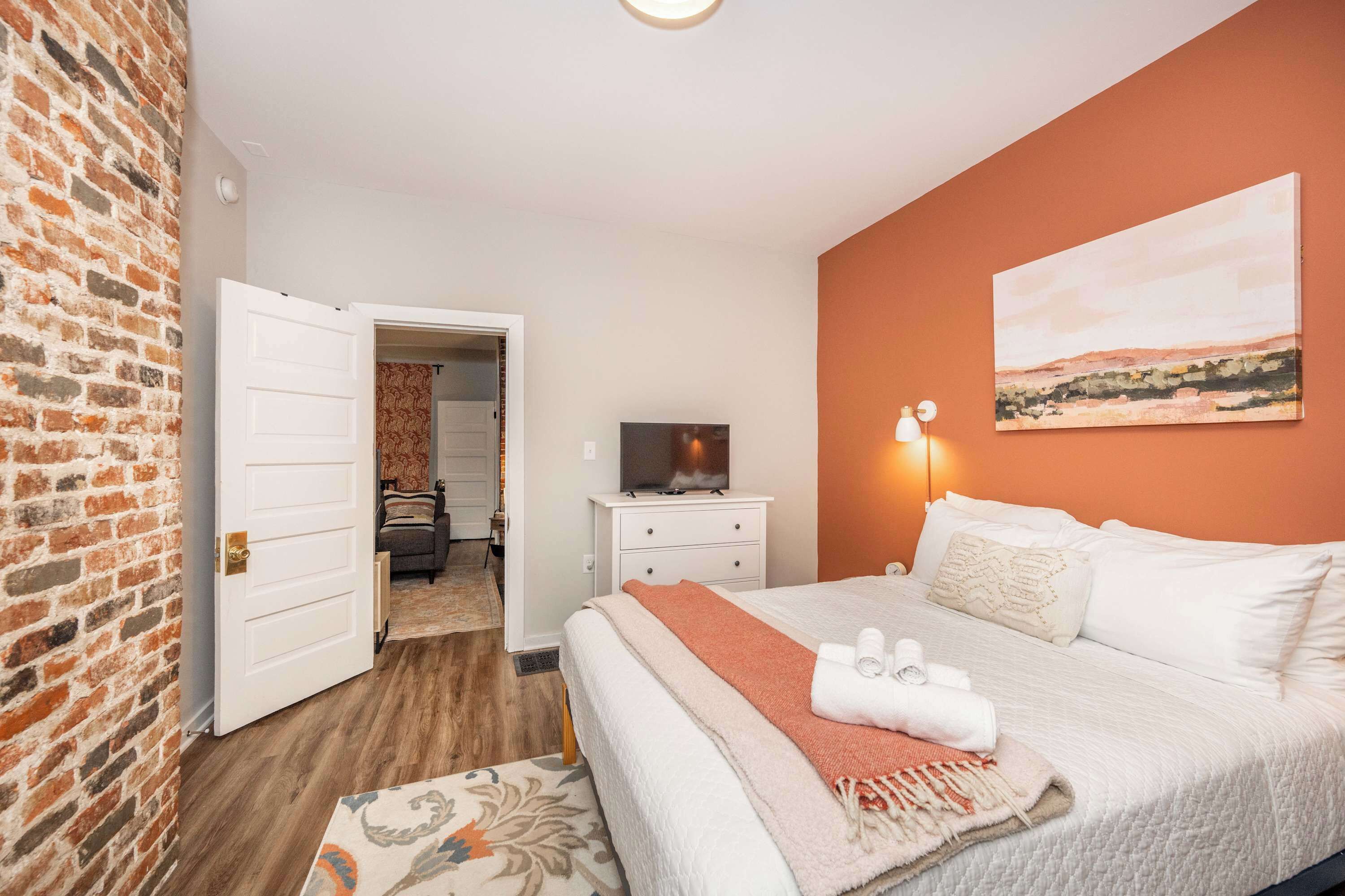 Une chambre d’hôtel avec un mur en briques, un lit avec une literie blanche, un téléviseur posé sur une commode blanche et un tableau accroché au mur.