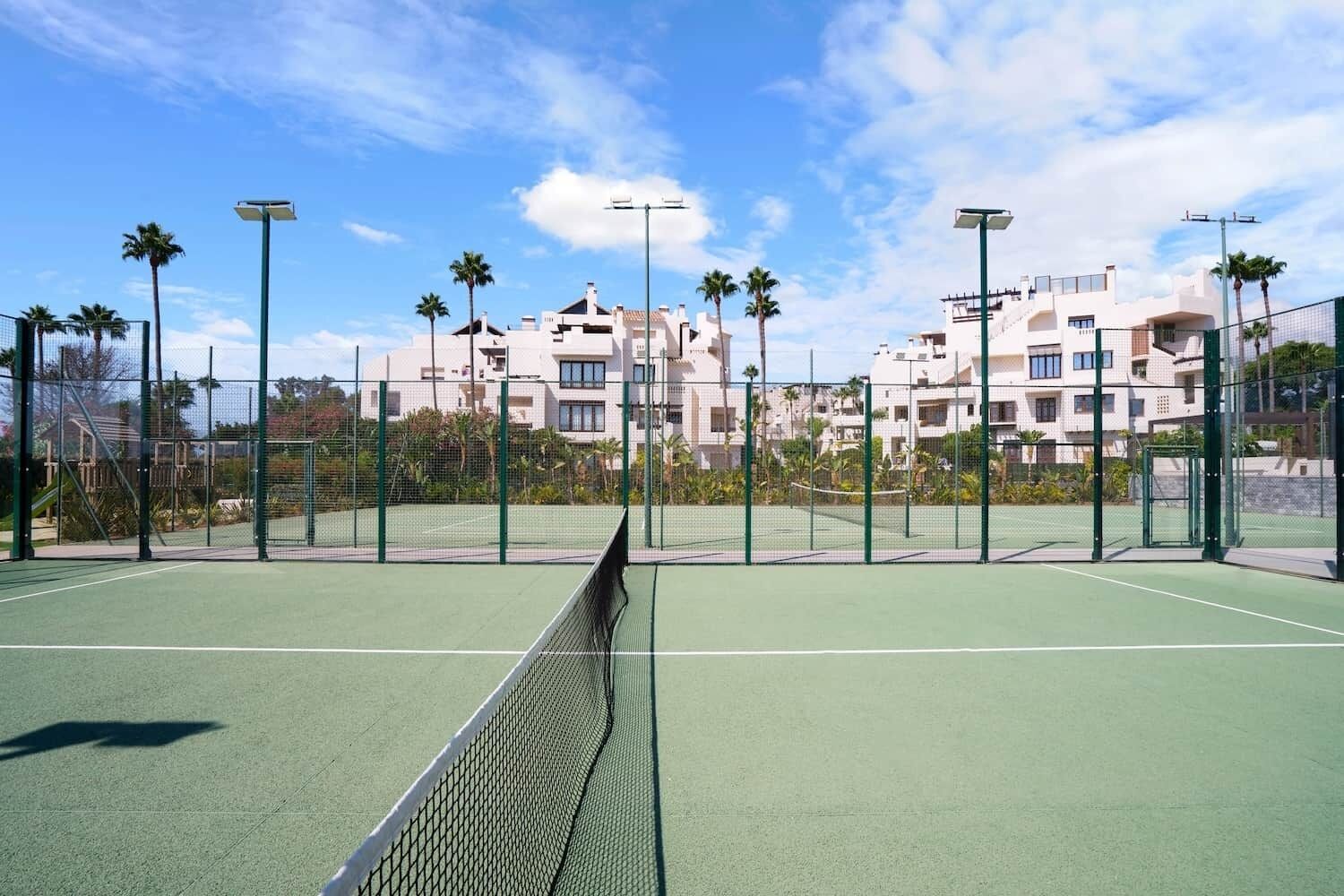 Un campo da tennis con una rete in primo piano, circondato da una recinzione e palme.