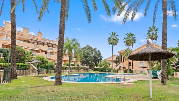 A pool area with palm trees, a thatched umbrella, and a building with balconies in the background. - RDR446 - Gorgeous 3 bedroom Santa Maria Golf (Marbella)