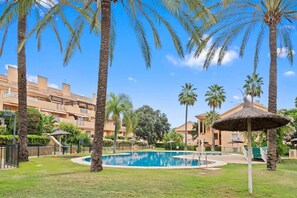 A pool area with palm trees, a thatched umbrella, and a building with balconies in the background. - RDR446 - Gorgeous 3 bedroom Santa Maria Golf (Marbella)