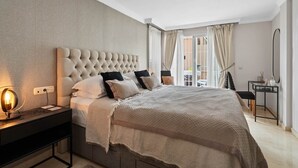 A modern bedroom with a tufted headboard, a large bed with patterned bedding, a bedside table with a lamp, and a view of the outdoors through a sliding glass door. - RDR446 - Gorgeous 3 bedroom Santa Maria Golf (Marbella)