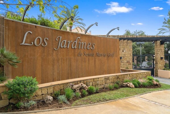 A wooden sign with "Los Jardines de Sagrada Maria Golf" written on it, surrounded by landscaped garden beds and a stone wall.