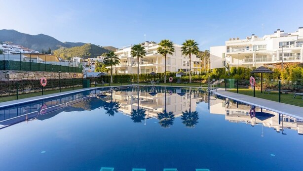 A clear swimming pool with a reflection of palm trees and buildings, surrounded by a fence and a grassy area. - RDR450 - Beautiful family apartment in Benahavis (Benahavís)