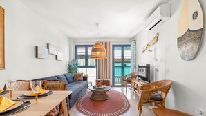 A modern living room with a sofa, dining table, and chairs. There is a TV, a plant, and a surfboard on the wall.