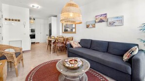 A modern living room with a grey sofa, wicker chairs, a glass coffee table with decorative stones, a hanging rattan light fixture, and a dining area with a white table and chairs.