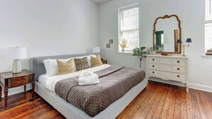 A bedroom with a bed, nightstands, a dresser, a mirror, and a window with blinds. - A State of Mind, Modern | Historic | Central (Savannah)