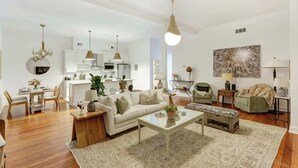 A modern living room with a sofa, coffee table, and a dining area with chairs and a chandelier. - A State of Mind, Modern | Historic | Central (Savannah)