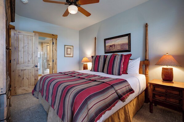 Une chambre à coucher avec un lit, une porte en bois, un ventilateur de plafond, une table de chevet avec une lampe et un tableau encadré au mur.