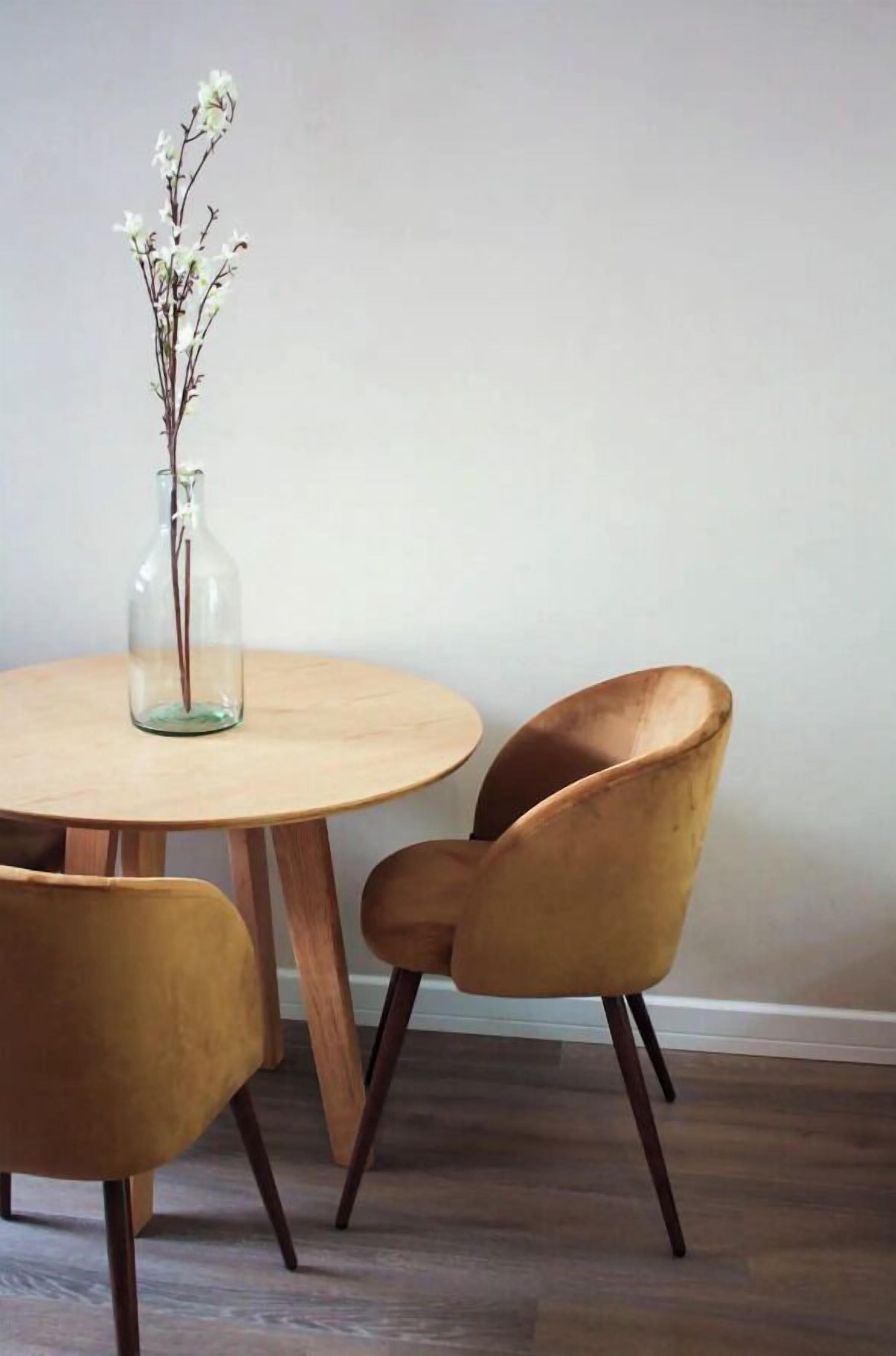 Een ronde houten tafel met twee stoelen, een glazen vaas met bloemen en een effen muur.