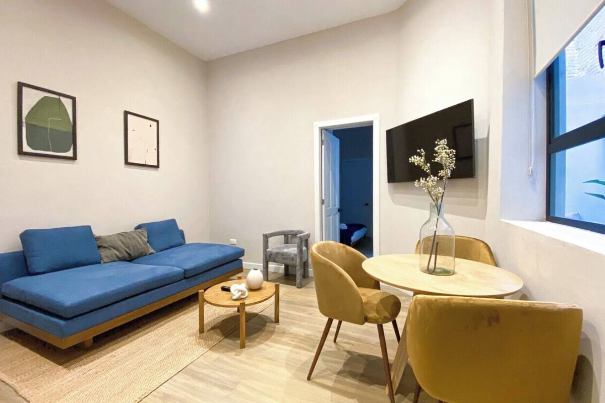A modern living room with a blue sofa, a round wooden table, and two yellow chairs. There is a small rug on the floor, and a vase with flowers on the table.