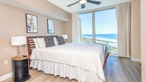 Habitación de hotel con una cama grande, ventilador de techo, mesita de noche con lámpara y vista a la playa por la ventana.