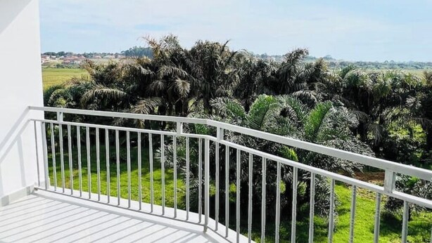 A balcony with a white railing overlooking a lush landscape with palm trees and a distant view of a town. - 1 Bedroom Apartment Palm-View Balcony Entebbe (Nkumba)