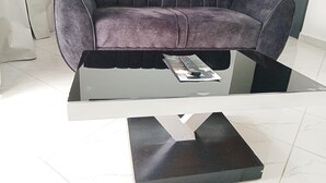 A modern living room with a dark grey sofa, a glass coffee table, and a remote control on the table. - 1 Bedroom Apartment Palm-View Balcony Entebbe (Nkumba)