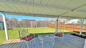 Un patio cubierto con un deck de madera, dos sillas de metal blanco con cojines rojos y una mesa de metal.