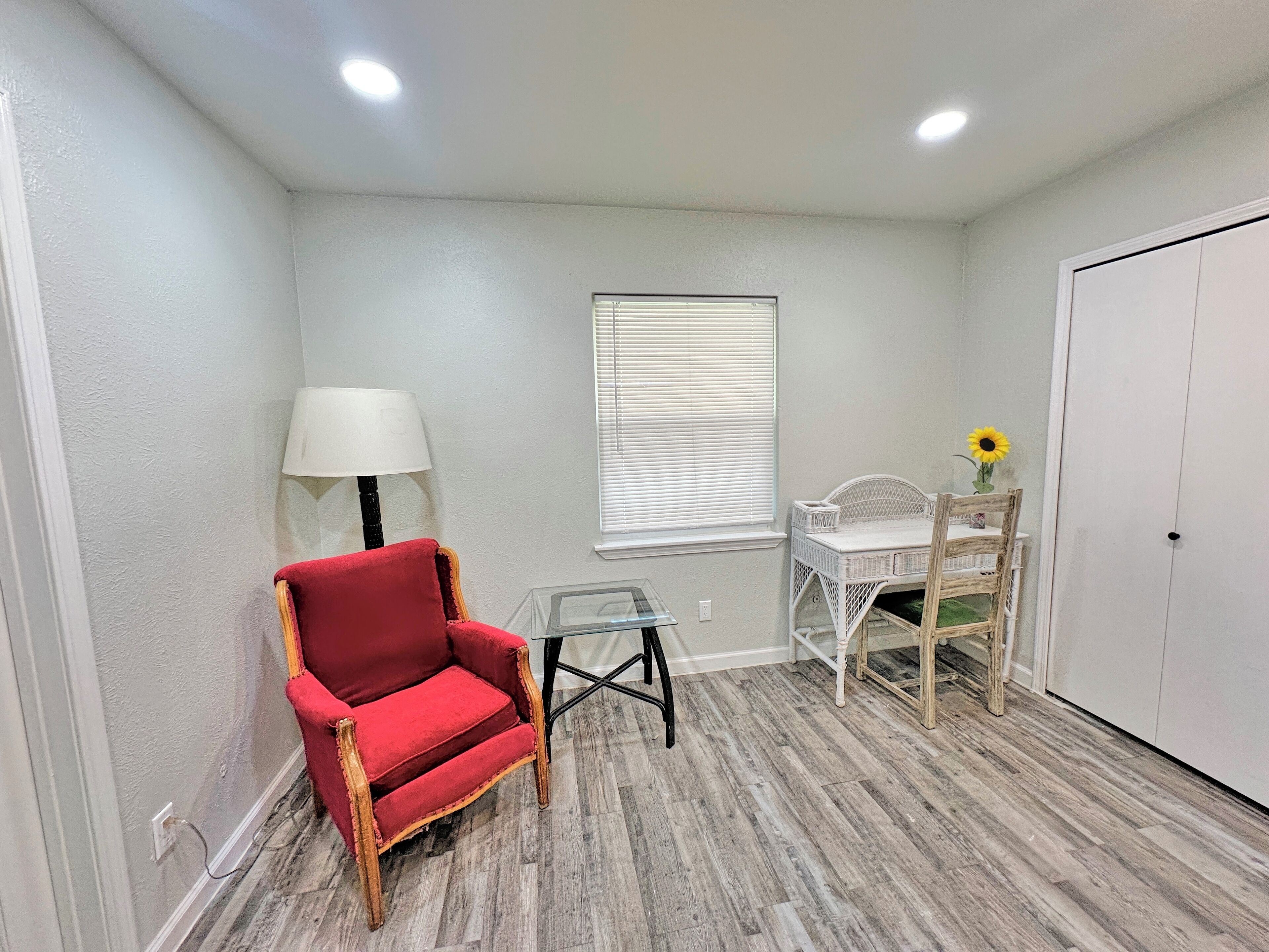 A room with a red chair, a small table, a lamp, a window with blinds, a wooden chair, and a door.