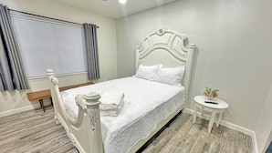 Un dormitorio con una cama blanca, un banco de madera, una mesita redonda con una planta y un ventilador de techo.