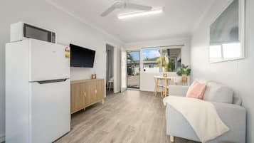 A compact kitchen with a refrigerator, microwave, and a TV. There's a dining area with a table and chairs, and a sofa with a cushion. The room has wooden flooring and white walls.