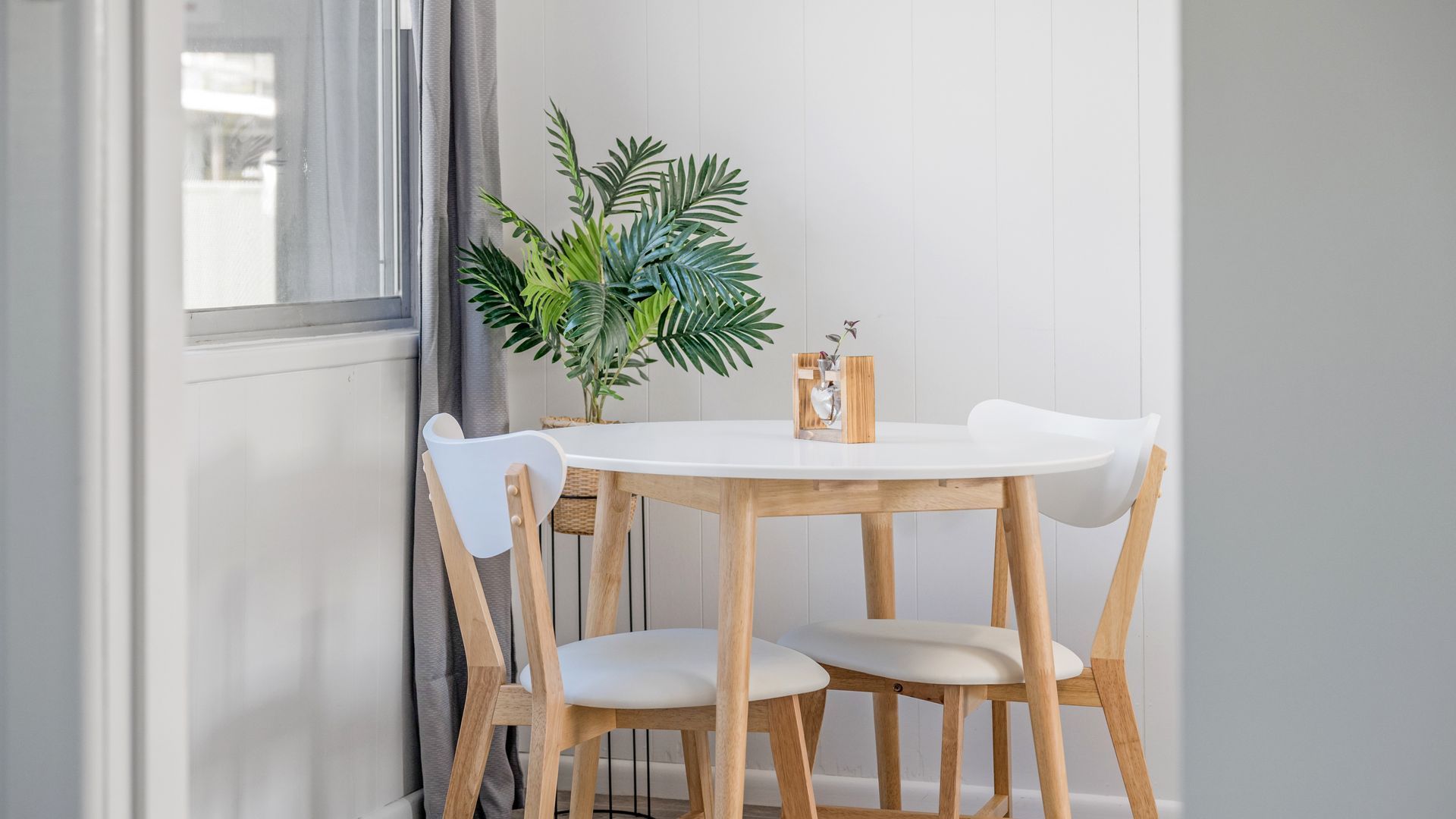 A round wooden table with four white chairs, a potted plant, and a small wooden box with a vase on it.