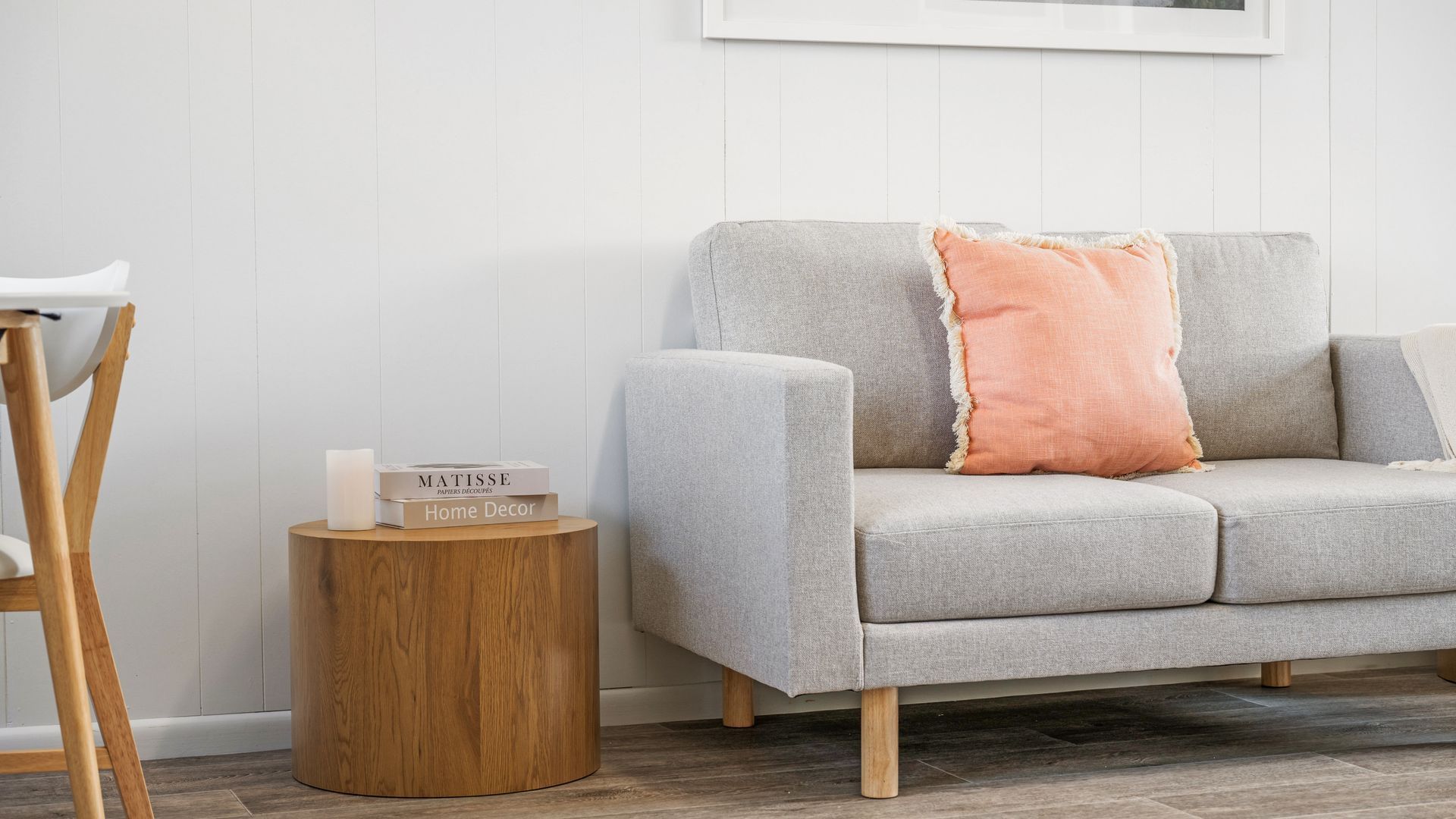 A neutral-toned sofa with a cushion, a small wooden side table with a book and candle, and a white wall with a framed picture.