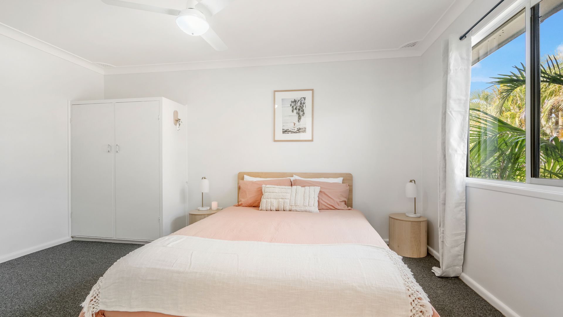 A bedroom with a bed, bedside tables, a wardrobe, a ceiling fan, a framed picture on the wall, and a window with a view of palm trees.