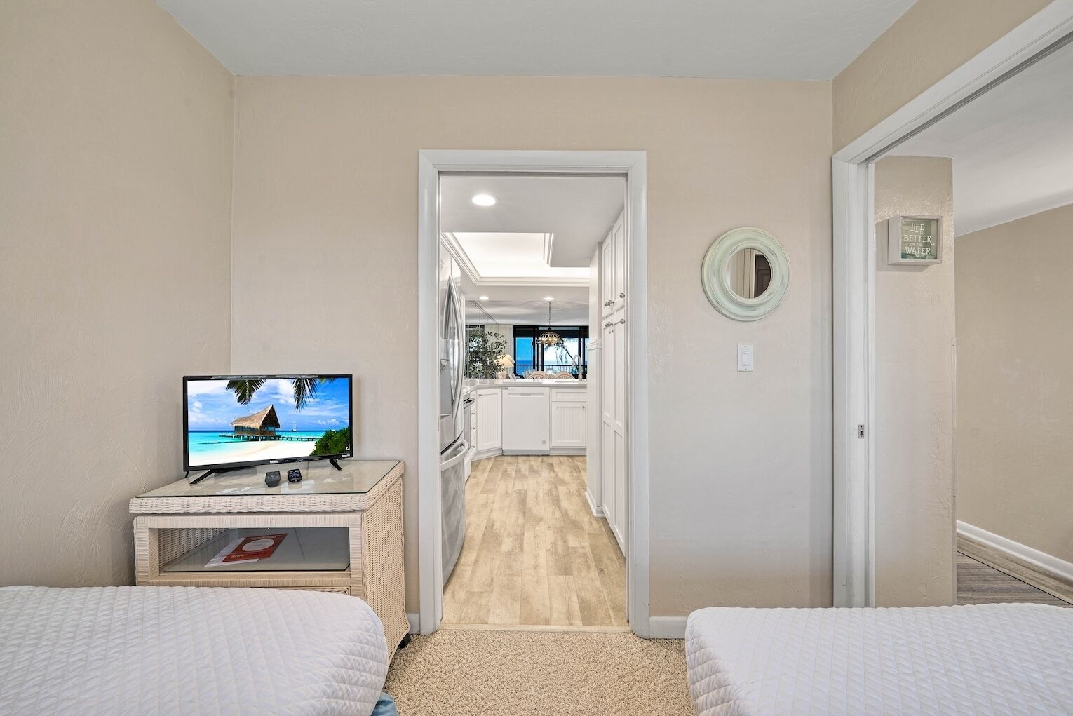 A hotel room with two beds, a TV on a side table, a kitchen area visible through an open door, and a mirror on the wall.