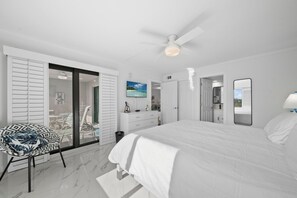 Un dormitorio moderno con una cama grande, ventilador de techo, televisor y baño con espejo y lavamanos.
