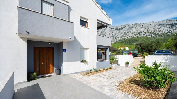A modern two-story building with a wooden door, a small outdoor seating area, and a view of a mountainous landscape. - Modern 2-Bedroom Apartment for up to 6 persons. (Orebic)