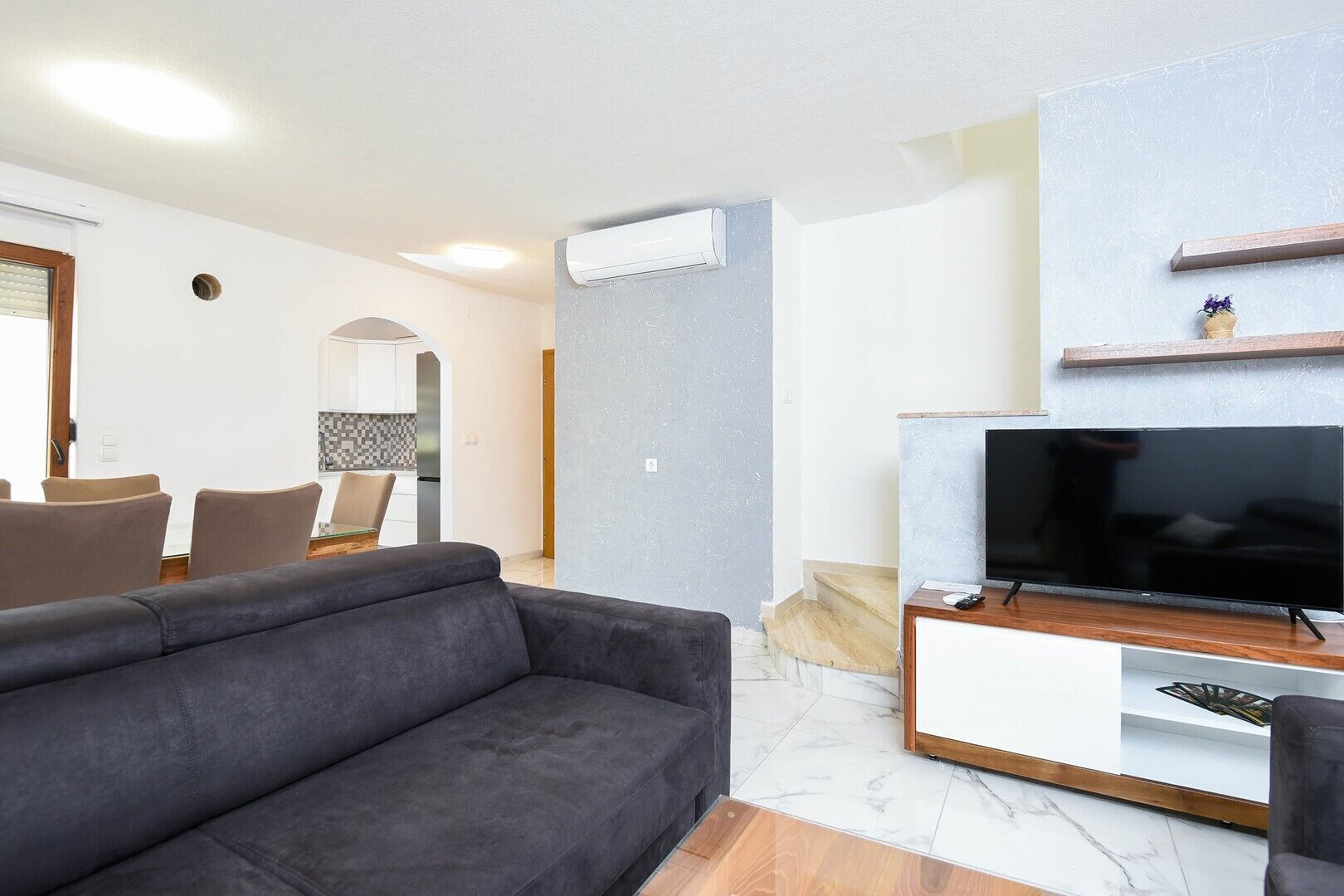 Una sala moderna con un sofá gris, un televisor de pantalla plana sobre un soporte de madera, y un comedor con sillas y un salpicadero de azulejos.