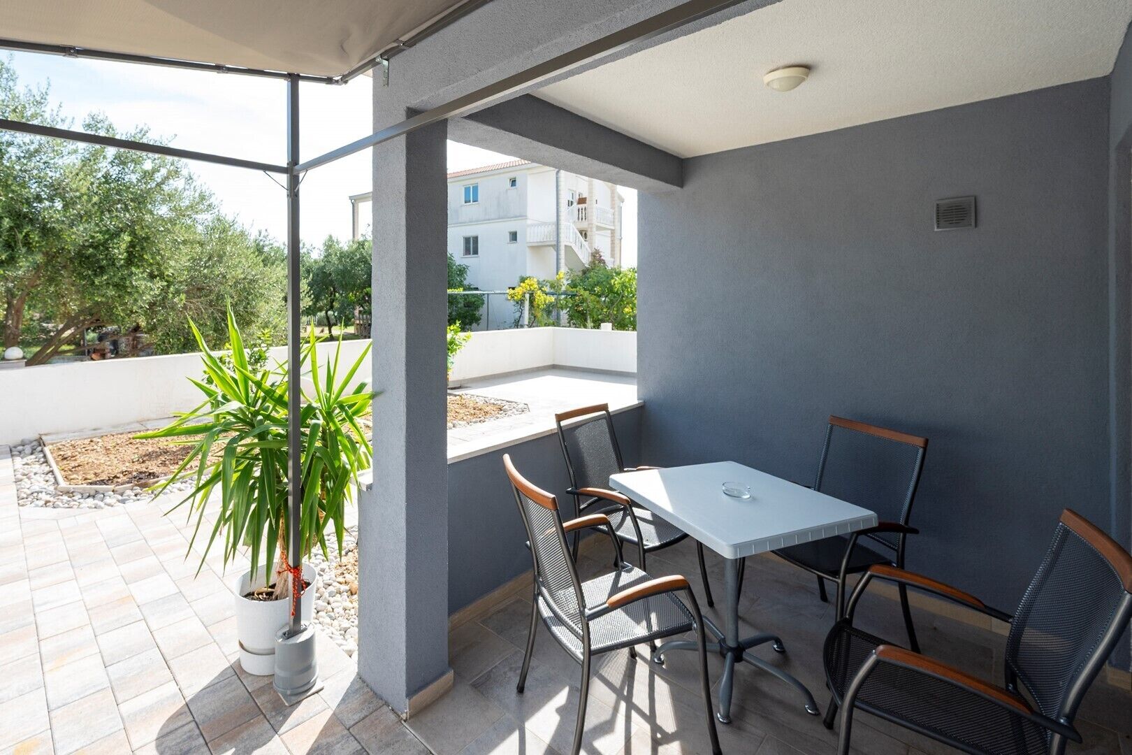A covered patio with a table and chairs, potted plants, and a view of buildings and trees.