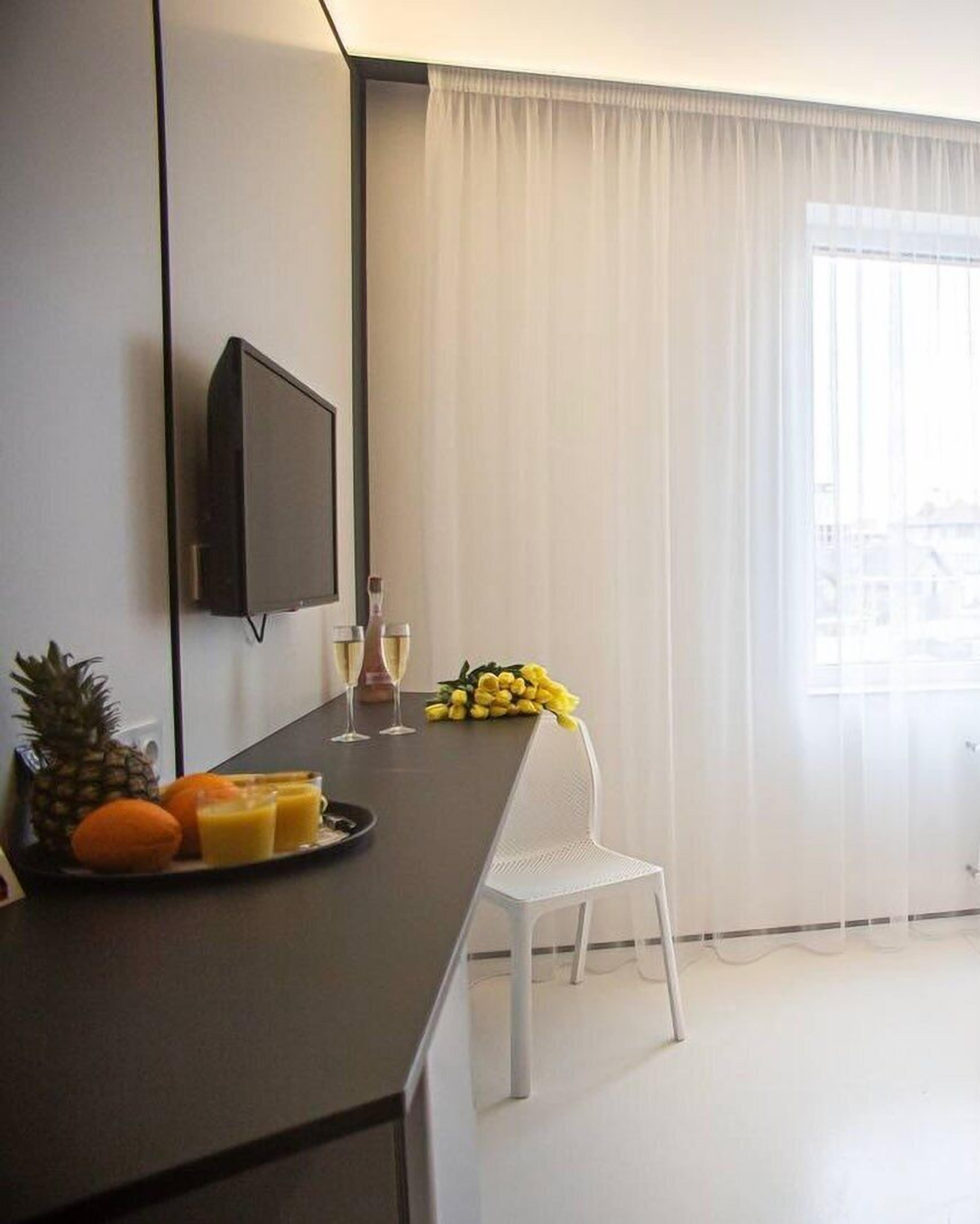 A hotel room with a dark-colored desk, a flat-screen TV mounted on the wall, a white chair, a dining table with fruits and glasses, and a window with sheer curtains.