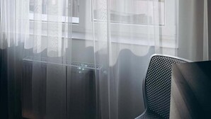 A room with sheer white curtains, a chair, and a view of tall buildings through the windows.