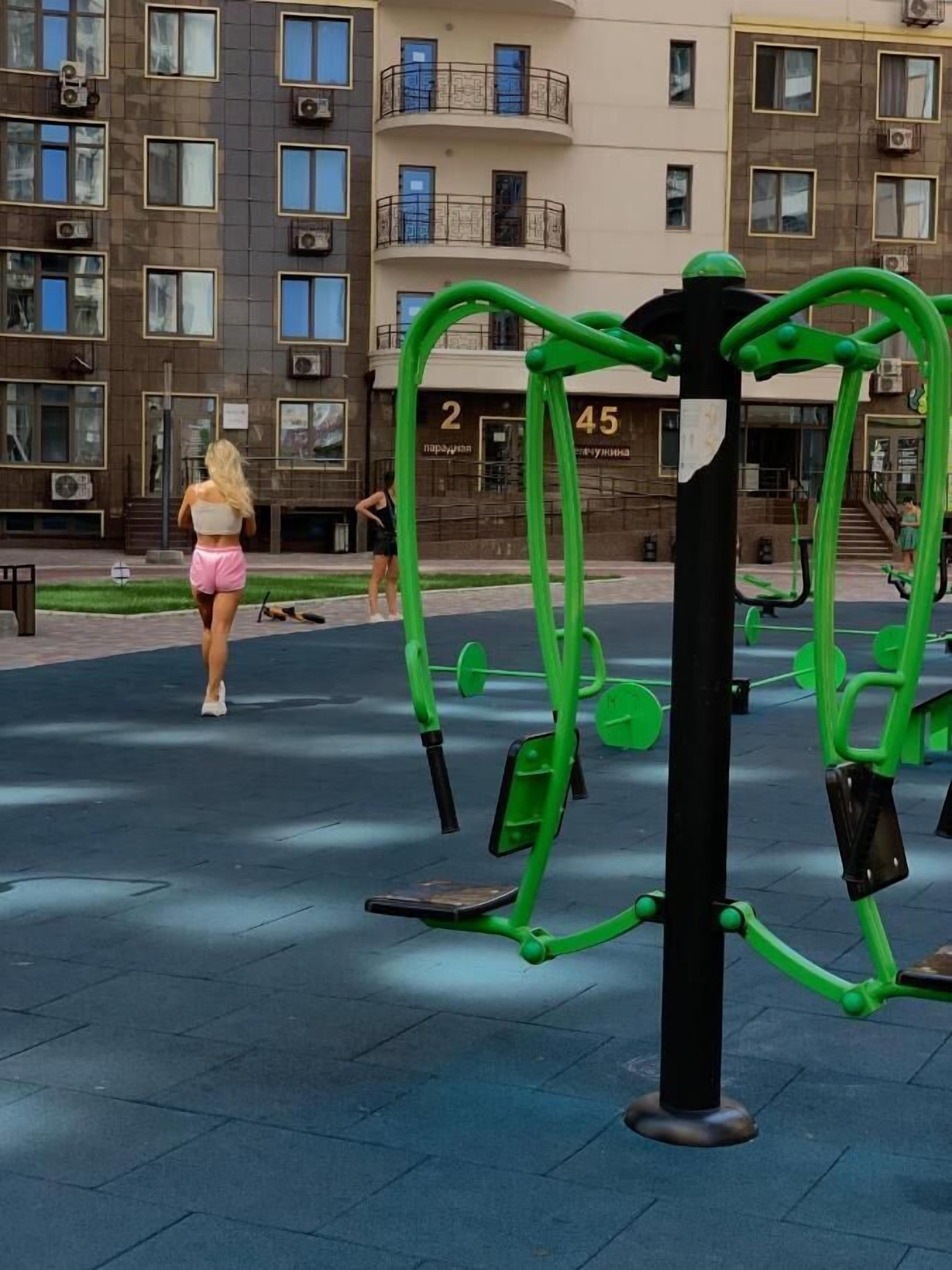 Een buiten trainingsgebied met groene fitnessapparatuur, een gebouw met balkons op de achtergrond, en een persoon die loopt.