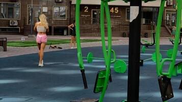 An outdoor exercise area with green fitness equipment, a building with balconies in the background, and a person walking.