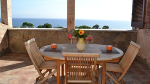 A round wooden table with four chairs, a vase with flowers, and two orange cups on a stone-paved patio overlooking the sea. - Villino Trilocale Apartments Seaside (Villaputzu)